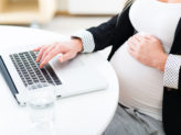 Pregnant women using a laptop on a white table with a cup of water next to her.