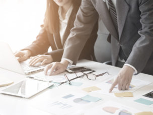 A business woman typing on a laptop with a business man pointing at a paper regarding a graphs they have analyzed.