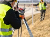 two men on walkie talkies and neon yellow safey vest on with yellow construction helmets with a surveyor theodolite on tripod