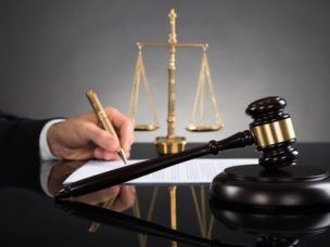 Close-up of judge writing on paper with a gavel and scale on the desk.