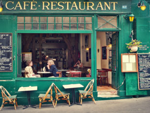 green cafe restaurant storefront with tables outside and two women sitting at table behind large open window