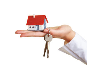 hand holding out a miniature house with a red roof and a set of silver keys